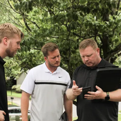 Three men discussing information on a tablet outdoors, surrounded by trees and greenery.