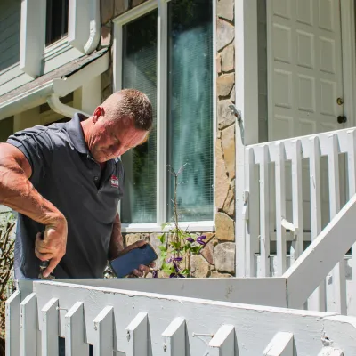 Man repairing white wooden railing on a house porch using a screwdriver on a sunny day.