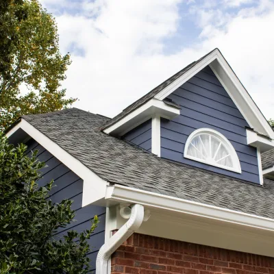 Close-up of a house roof with gray shingles, blue siding, white trim, and an arched window under a cloudy sky.