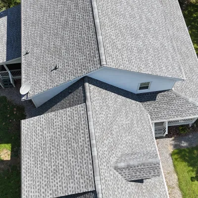 Aerial view of a grey shingled roof on a white house surrounded by green grass and trees on a sunny day.