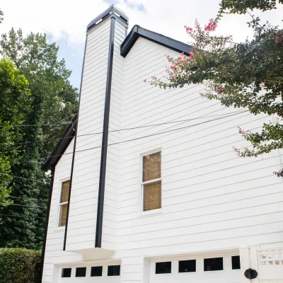 White two-story house with black trim, tall chimney, and surrounding green trees under a cloudy sky