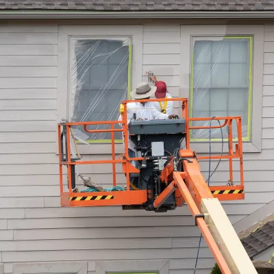 Two workers using a lift to paint the exterior of a house with partially covered windows.