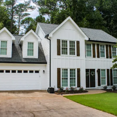 Two-story white house with gray roof, double garage, wooden shutters, and green lawn surrounded by trees.