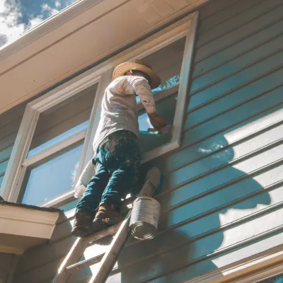 A painter applying blue paint to a house window while standing on a ladder under a sunny sky.