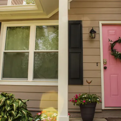 Pink front door with green wreath on beige house exterior with window, black shutters, and potted plants.