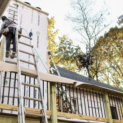 A worker installs siding on a home, using ladders and tools against a backdrop of autumn trees.