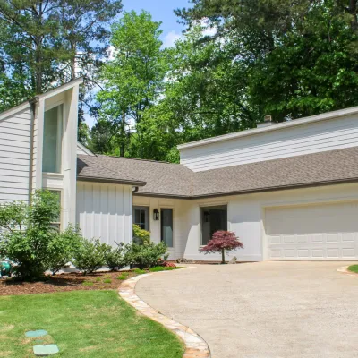Modern single-story house with white siding, a two-car garage, and a curved driveway surrounded by greenery.