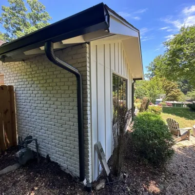 Side of white brick and siding house with black gutter, garden area, wooden fence, and sunny backyard with chairs and basketball hoop.