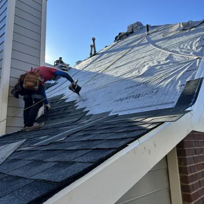 Roofer installing shingles on a sloped roof under clear blue sky during daytime construction work