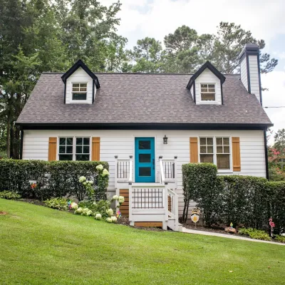 Charming white cottage with blue door, dormer windows, and manicured bushes on a green lawn under a cloudy sky.