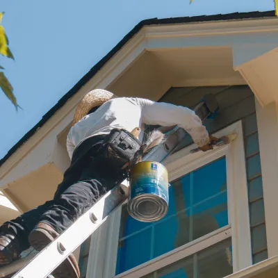 A painter on a ladder applying paint to a window frame on a house under bright blue skies.