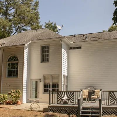 Backyard view of a two-story white house with a deck, outdoor seating, and trees surrounding.