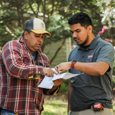 Two men outdoors discussing and pointing at a document, with trees and a house in the background.