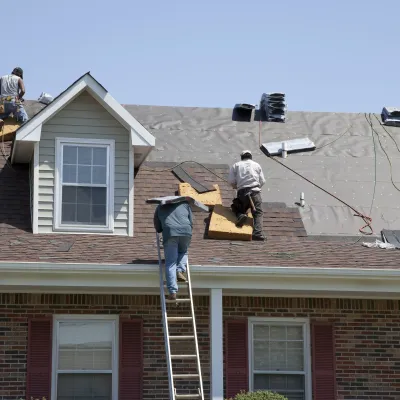 Workers repairing a residential roof with tools, ladders, and materials under clear blue skies.