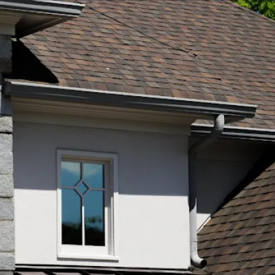 House exterior showing stone wall, white framed window, and brown shingled roof with gutter and blue sky.