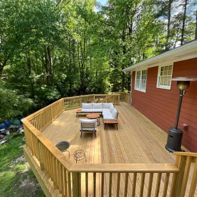 Spacious wooden deck with outdoor seating set and patio heater beside red house surrounded by green trees.