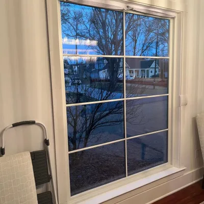 Large window with grid pattern showing outdoor tree and neighborhood at dusk inside a cozy room with chairs and ladder.
