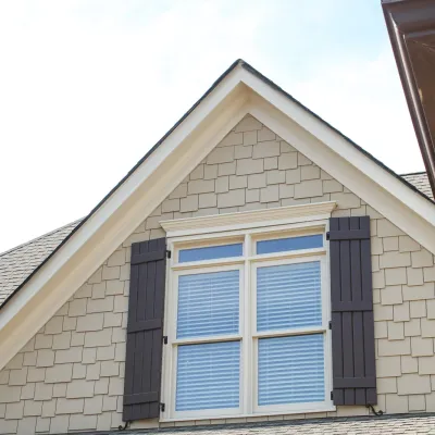 Close-up of a beige shingle house facade with white window frame and dark shutters under blue sky.