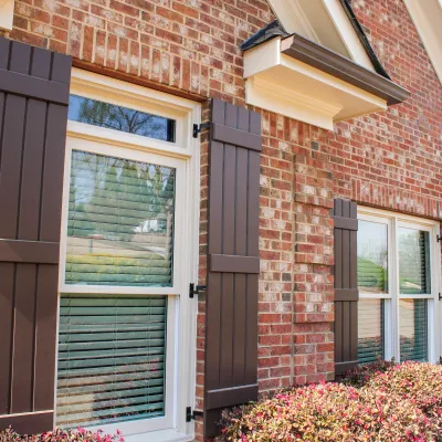 Red brick house exterior with brown window shutters and bushes under the windows on a sunny day