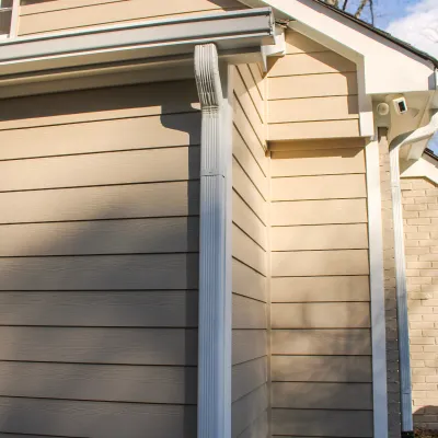 Close-up of beige house exterior with white gutters and drainpipes under sunlight in a suburban setting.