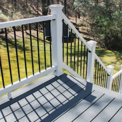 White railing with black balusters on a sunlit gray deck overlooking grassy backyard and trees.