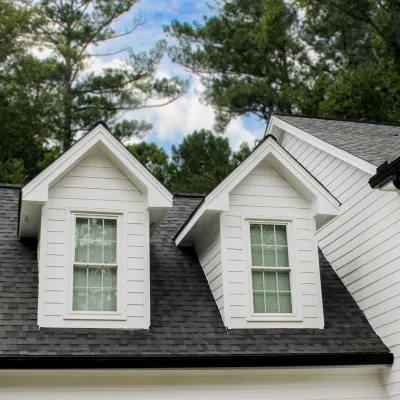 White house dormer windows with black roof shingles surrounded by green trees under blue sky.
