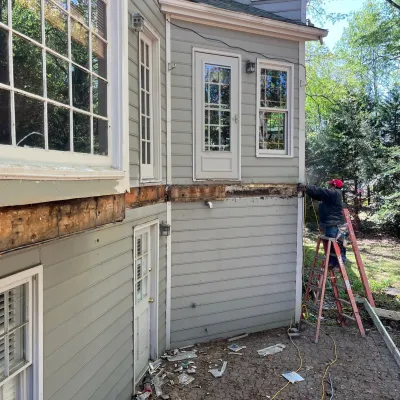 Worker repairing exterior siding on a gray two-story house using a ladder surrounded by tools and debris.