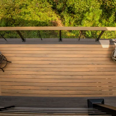 A wooden balcony with a round table, three chairs, and a reclining chair overlooking lush greenery.