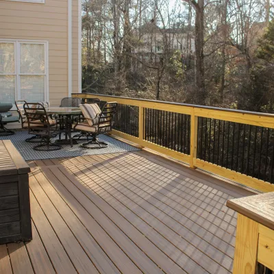 Sunny wooden deck with outdoor seating area, metal railing, storage bench, and trees in the background.