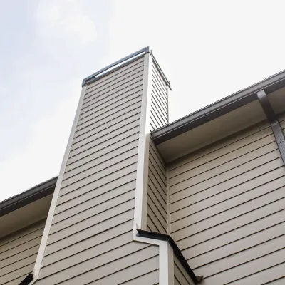 Close-up of beige siding on a house corner with a chimney and roof against a cloudy sky