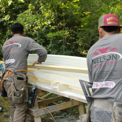 Two construction workers handling wooden planks outdoors with tools and safety gear.