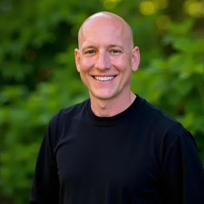 Smiling bald man wearing a black shirt standing outdoors with green leafy background in soft focus