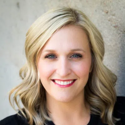 Portrait of a smiling blonde woman with blue eyes against a neutral textured background.