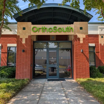 Entrance of OrthoSouth clinic with green signage, red brick walls, glass door, and surrounding greenery under blue sky.