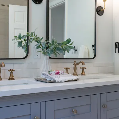 Modern bathroom vanity with double sinks, brass faucets, rectangular mirrors, and decorative greenery in white vase.