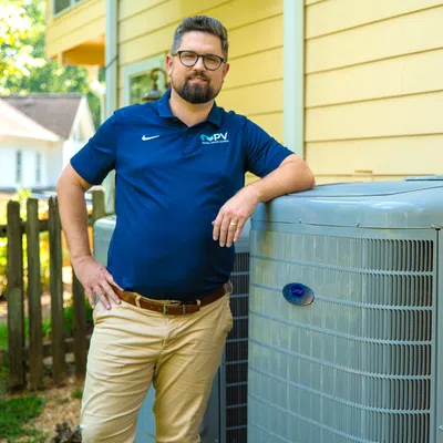 Technician in blue shirt posing with a modern air conditioning unit outside a yellow house on a sunny day.
