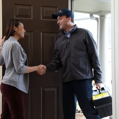 A service technician greets a homeowner at the front door, offering assistance and smiling.