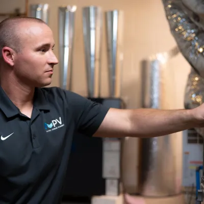 Technician in PV uniform adjusting HVAC system components in a utility room with metal ducts and pipes.