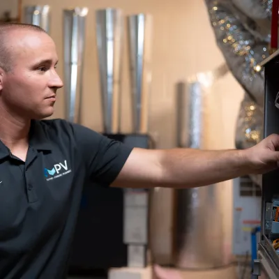 Technician in PV uniform adjusting HVAC system components in a utility room with metal ducts and pipes.