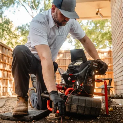 Technician using a RIDGID SeeSnake inspection camera to inspect an outdoor drainage pipe in a backyard.