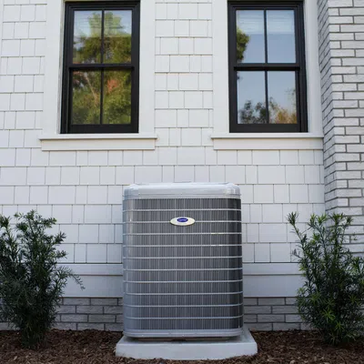 Outdoor air conditioning unit installed against white shingle and brick house wall with plants on both sides