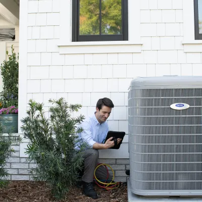 Technician inspecting and testing a Carrier air conditioning unit outside a modern house with white siding.