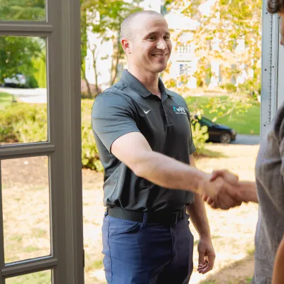 Two men shaking hands at a doorway, one in a black polo and the other in a gray shirt, outdoors in daylight