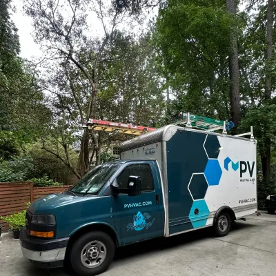 Blue and white PV Heating & Air plumbing service van parked in a wooded residential driveway with ladders on top.
