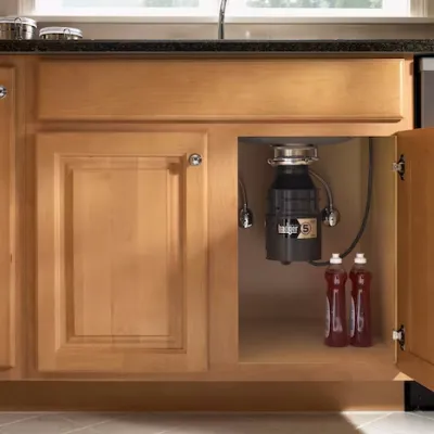 Kitchen cabinet with open door showing installed garbage disposal and two red liquid bottles inside.