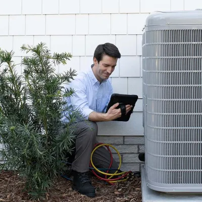 Technician using a tablet to inspect an outdoor air conditioning unit beside a house exterior with plants.