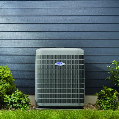 Outdoor air conditioning unit installed against blue siding wall with shrubs and grass around it