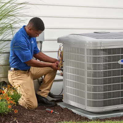 Technician in blue shirt repairing air conditioning unit outside a white house near garden flowers.