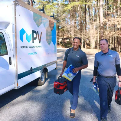 Two HVAC technicians carrying toolboxes walking away from PV heating and cooling service van on sunny day.