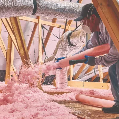 Worker in protective gear installing pink fiberglass insulation in attic space with wooden beams and ducts.